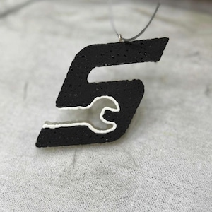 May include: A black pendant in the shape of the letter "S" with a white wrench design inside. The pendant is suspended from a thin, silver-colored cord. The background is a light gray, textured surface.