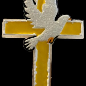 May include: A decorative cross with a yellow center and white border, featuring a white dove with glitter accents. The dove is positioned over the cross, with its wings spread. The background is black.