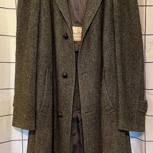 May include: A long, dark olive green and brown tweed overcoat featuring a notched lapel, three dark buttons down the front, and long sleeves with buttoned cuffs. The interior is lined with a dark fabric and includes a visible Harris Tweed label.