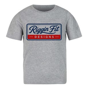 May include: A heather gray short-sleeve t-shirt featuring a rectangular logo on the chest. The logo has a dark blue script text that reads Riggin Fit above a red horizontal bar containing the white text DESIGNS, all enclosed in a dark blue border.