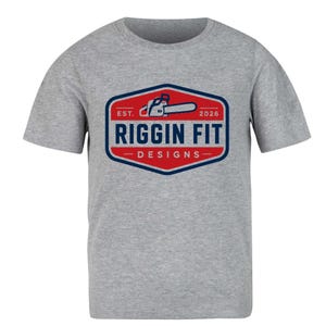May include: A heather gray short-sleeve t-shirt featuring a central graphic of a red and blue hexagon. Inside the hexagon is a chainsaw icon, the text EST. 2026, the words RIGGIN FIT in large blue letters, and the word DESIGNS in smaller blue letters.