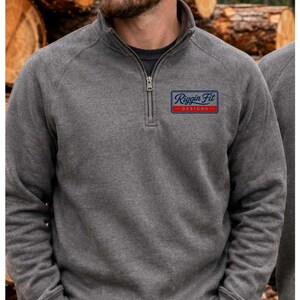 May include: A charcoal gray quarter-zip sweatshirt featuring a rectangular patch on the left chest with the text Riggin Fit DESIGNS in blue and red. The sweatshirt has long sleeves with ribbed cuffs and a high collar with a silver metal zipper.
