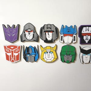 Può includere: Un set di dieci magneti piatti sagomati che raffigurano volti di personaggi e simboli stilizzati della serie Transformers. Il set include il logo viola dei Decepticon, il logo rosso degli Autobot e varie teste di robot in blu, giallo, verde e grigio.