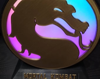 Könnte beinhalten: Ein 3D-gedrucktes, goldfarbenes Mortal Kombat-Logo mit schwarzem Sockel. Das Logo wird mit einem Regenbogen aus Farben beleuchtet, darunter Lila, Blau und Grün.