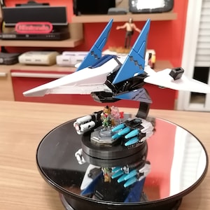Peut inclure: Un modèle de vaisseau spatial blanc et bleu avec une base noire et une plateforme rotative. Le vaisseau spatial a une pointe jaune et des ailes bleues. La base a une petite figurine d'un personnage de jeu vidéo.