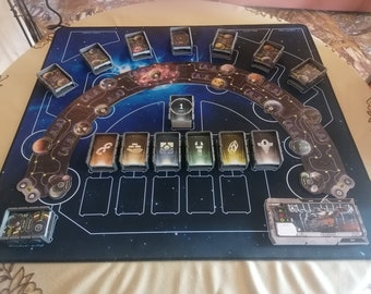 Könnte beinhalten: Ein schwarzes und blaues Spielbrett mit einem Galaxie-Design. Das Brett hat eine kreisförmige Bahn mit Planeten und Sternen. Es gibt mehrere kleine Kartenhalter mit Karten darin. Das Brett hat auch einen zentralen Bereich mit einem Token mit der Nummer 1 und mehreren anderen Kartenhaltern. Das Brett ist aus einem dicken, strapazierfähigen Material gefertigt.
