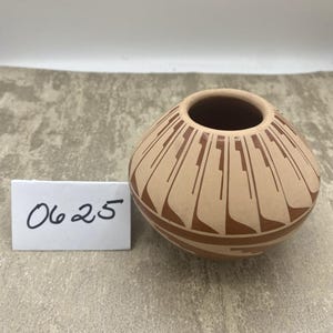 May include: A small, round, tan-colored ceramic pot featuring a dark brown geometric pattern radiating from the opening. A white rectangular card with the handwritten black text 0625 is positioned to the left of the ceramic vessel.