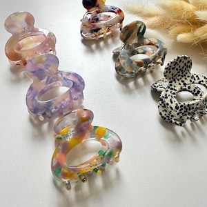 Könnte beinhalten: Eine Sammlung farbenfroher Haarspangen mit verschiedenen Mustern. Die Spangen weisen ein marmoriertes Design mit Rosa-, Lila-, Gelb- und Grüntönen auf. Eine Spange hat ein schwarz-weißes gepunktetes Muster. Die Spangen bestehen aus glänzendem Material.