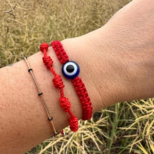 Könnte beinhalten: Drei Armbänder werden an einem Handgelenk gezeigt. Eines ist goldfarben mit schwarzen Perlen, das zweite ist rot mit Knoten und das dritte ist ein rot gewebtes Armband mit einer blau-weißen Evil-Eye-Perle. Die Armbänder werden zusammen getragen.