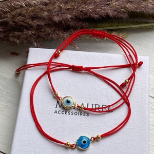 Könnte beinhalten: Zwei rote Armbänder mit goldenen Perlen und blauen bösen Augen-Anhängern. Die Armbänder liegen auf einer weißen Schachtel mit dem Aufdruck "ACCESSOIRES".
