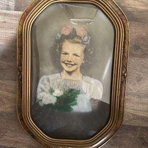 May include: A vintage portrait of a smiling child wearing a floral headpiece and a light-colored dress, holding a bouquet of white flowers. The portrait is encased in an ornate, oval-shaped gold-toned frame with a convex glass cover.