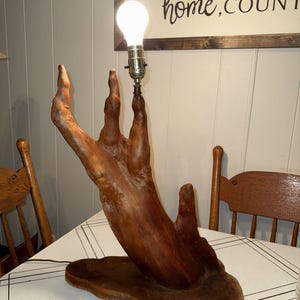 May include: A table lamp featuring a base made from a natural, reddish-brown piece of driftwood shaped like an upright hand with extended fingers. A silver-toned metal light socket is attached to the tip of the middle finger, holding a glowing bulb.