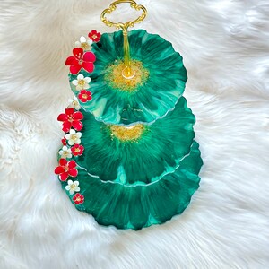 Könnte beinhalten: Dreistöckiger Serviertablett mit einem goldenen Metallständer. Jede Etage ist ein grüner und weißer Harz-Kreis mit goldenen Glitzerakzenten. Die oberste Etage ist mit roten und weißen Blumen verziert.