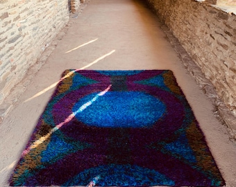 May include: A rectangular, shaggy rug with a vibrant, abstract design. The rug features a central blue oval shape, surrounded by purple, teal, and gold accents. The rug is placed on a concrete surface, with brick walls in the background.