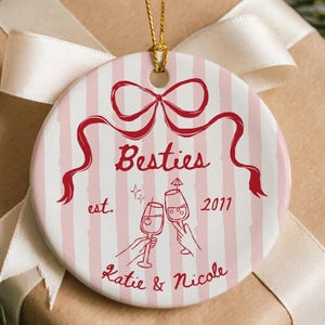 Puede incluir: Adorno navideño redondo, a rayas rosas y blancas, con la palabra "Besties" en rojo, un lazo y el texto "est. 2011". Dos manos brindan con copas de vino. Los nombres "Katie & Nicole" están escritos debajo.