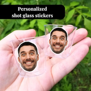 May include: Two clear plastic shot glasses with personalized face stickers. The stickers feature a smiling man's face.