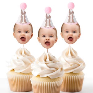 May include: Three cupcakes with pink and white bunny-themed cupcake toppers. Each topper features a baby's face with a pink and white hat with the number "1" on it. The cupcakes are frosted with white icing.