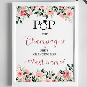 May include: A white sign with pink floral borders and the text "Pop the Champagne She's Changing Her Last Name!"