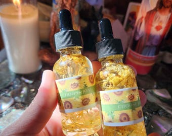 May include: Two small glass bottles with black droppers, filled with yellow liquid and flower petals. Each bottle has a label with sunflowers and the text "CHOOSE JOY!". The bottles are held in a hand.