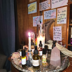 May include: A collection of essential oil bottles, candles, crystals, and inspirational notes arranged on a decorative table. The scene includes a lit candle, a rose quartz crystal, and a sign that says "Hocus Pocus".