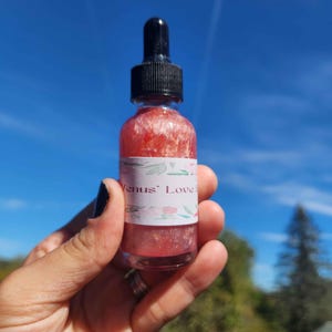 May include: A small glass bottle with a black dropper top, filled with a shimmering pink liquid. The bottle has a white label with the text "Venus' Love" and floral accents. The background is a clear blue sky.