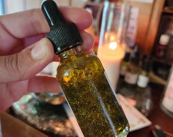 May include: A small glass bottle with a black dropper filled with a golden liquid containing glitter and small particles. The bottle is held in a hand, with a blurred background of candles and other items.
