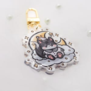 Könnte beinhalten: Ein Schlüsselanhänger aus klarem Acryl mit einem Cartoon-Wolf in Schlafmütze, der auf einer grauen Wolke vor einem gelben Halbmond und Sternen sitzt. Der Anhänger ist an einem goldfarbenen Karabinerverschluss aus Metall befestigt.