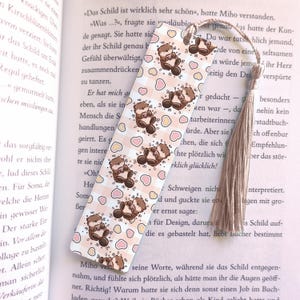 Op de afbeelding: Decoratieve boekenlegger met een herhaald patroon van cartoonotters en harten op een lichtroze en witte geruite achtergrond. De boekenlegger heeft een beige kwast. Voor boekenliefhebbers.