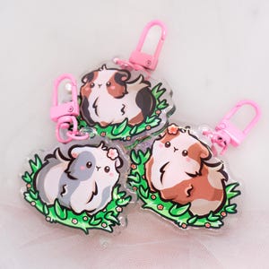 Könnte beinhalten: Drei transparente Acryl-Schlüsselanhänger mit Cartoon-Meerschweinchen in den Farben Braun, Weiß und Grau. Jeder Schlüsselanhänger hat einen rosa Verschluss und ist von grünen Blättern und kleinen rosa Blüten umgeben.
