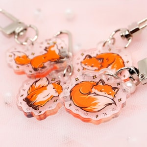 Könnte beinhalten: Vier transparente Acryl-Schlüsselanhänger mit orangefarbenen Fuchs-Illustrationen. Jeder Schlüsselanhänger zeigt eine andere Pose des Fuchses, darunter schlafend, stehend und zusammengerollt. Die Schlüsselanhänger haben einen silbernen Metallring und einen kleinen Perlenanhänger.