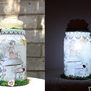Op de afbeelding: Een glazen pot versierd met een fee die op een paddenstoel zit, omringd door bloemen en lieveheersbeestjes. De pot heeft een witte kantversiering en een witte bloem bovenop. De pot wordt van binnenuit verlicht, waardoor een warme gloed ontstaat.