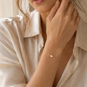May include: A delicate gold chain bracelet featuring a single small white oval pearl centered on the wrist. The bracelet is constructed with a thin metallic link chain and a secure clasp, showcasing a minimalist design that rests against the skin.