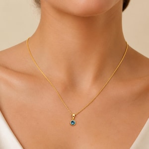 Puede incluir: Un collar de cadena dorada con un colgante de piedra preciosa azul redonda engastada en un bisel dorado. El colgante cuelga centralmente de una delicada cadena de eslabones dorados, descansando sobre la zona de la clavícula.