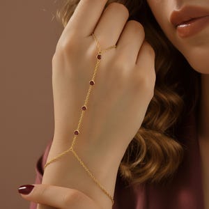 May include: A gold hand chain with a ring and bracelet connection, featuring a delicate chain design. The chain is adorned with five small, round, red gemstones. The jewelry is worn on a hand with a burgundy nail polish.