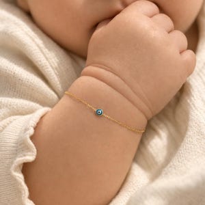 Könnte beinhalten: Ein zartes goldenes Armband mit einem kleinen, runden, blau-weißen Evil-Eye-Anhänger. Das Armband befindet sich am Handgelenk eines Babys, mit der Hand des Babys in der Nähe seines Mundes. Der Hintergrund ist ein weicher, cremefarbener Stoff.