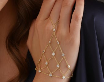 Op de afbeelding: Een goudkleurige handkettingarmband met een ruitvormig netontwerp. De armband heeft kleine, heldere edelstenen op de kruispunten van het net en een delicate ketting die verbonden is met een ring.
