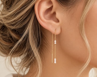 May include: A gold threader earring featuring a delicate chain that passes through the earlobe. The earring includes a straight metal bar at one end and two small clusters of white pearls spaced along the chain, creating a dangling effect.