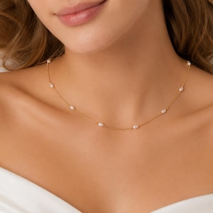 Puede incluir: Un collar de cadena dorada delicada con pequeñas perlas blancas ovaladas espaciadas uniformemente. El collar descansa sobre la clavícula, mostrando un diseño minimalista con una cadena de eslabones metálicos finos y varias perlas individuales.