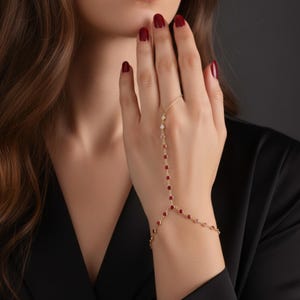 Może przedstawiać: Złoty łańcuszek na dłoń z czerwonymi i białymi kamieniami szlachetnymi. Łańcuszek łączy się z pierścionkiem i owija wokół nadgarstka. Kamienie szlachetne są małe i okrągłe, osadzone w delikatnym złotym łańcuszku.