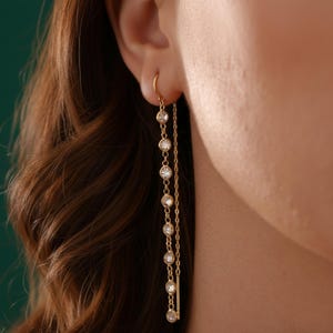 May include: Gold-toned drop earrings featuring a series of clear, round gemstones. The earrings have a delicate chain design, with the gemstones evenly spaced along the length. The earrings are shown against a blurred background.