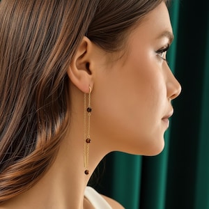 Peut inclure: Boucles d'oreilles pendantes dorées avec trois perles marron foncé suspendues à de fines chaînes. Les boucles d'oreilles ont un design simple et élégant avec un petit élément en forme de barre. Les boucles d'oreilles sont portées.