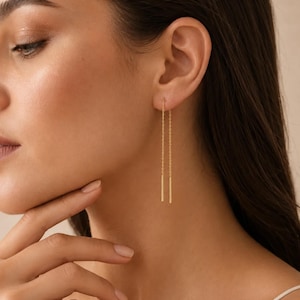 Peut inclure: Une paire de boucles d'oreilles en fil d'or composée d'une fine chaîne délicate qui traverse le lobe de l'oreille, se terminant par une fine barre dorée polie à chaque extrémité. Le design minimaliste pend verticalement avec élégance.