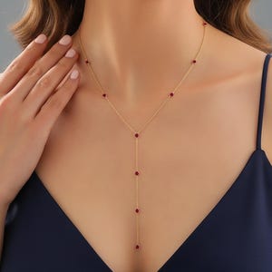Puede incluir: Un delicado collar dorado con un colgante en forma de Y. El collar presenta pequeñas piedras preciosas rojas redondas espaciadas uniformemente a lo largo de la cadena y el colgante. El collar se muestra sobre un fondo azul oscuro.