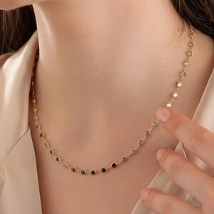 Puede incluir: Un delicado collar de oro con una serie de pequeñas piedras preciosas de colores. Las piedras preciosas son redondas y varían en color, incluyendo azul, verde y transparente. El collar se lleva alrededor del cuello.