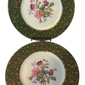 May include: Two decorative plates with a white center featuring a floral design of pink roses, purple flowers, and green leaves. The plates have a dark green border with a gold floral pattern and a gold rim.