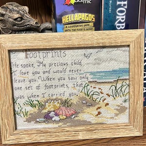 May include: A framed cross-stitch artwork with the text "Footprints" and a quote about love and support. The artwork depicts a beach scene with footprints in the sand, a seashell, and a parasol. The frame is made of light-colored wood.