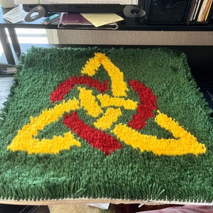 May include: Handmade green rug with a Celtic knot design in yellow and red. The rug is square-shaped and has a textured, shaggy appearance. The knot is centered on the rug, creating a striking visual contrast against the green background.
