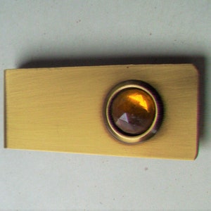 May include: A rectangular gold-toned metal money clip with a brushed finish. It features a circular, faceted amber-colored stone set in a dark metal bezel. The clip is designed to hold folded bills.