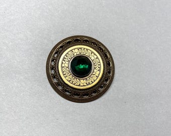 May include: A round, antique-style brooch with a central emerald-green gemstone. The brooch features a gold-colored inner circle with ornate floral detailing, surrounded by a bronze-colored filigree border. The brooch is a vintage accessory.