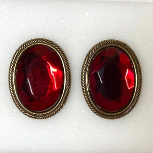 May include: A pair of oval-shaped earrings with a red gemstone set in a gold-toned metal frame.